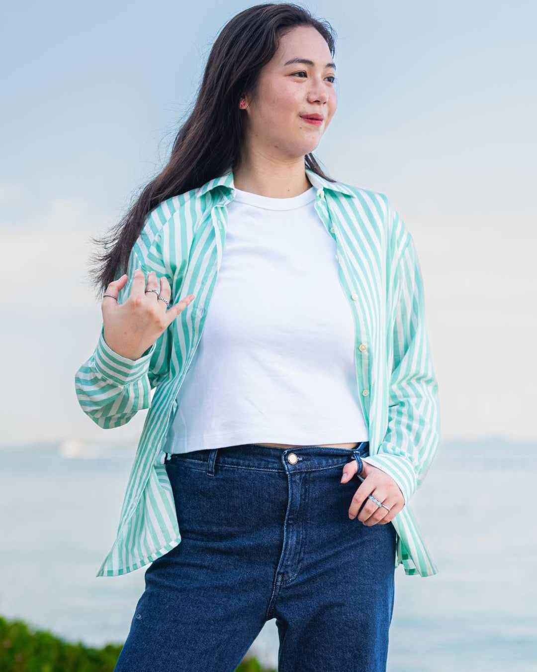 Woman wearing a green and white striped shirt over a white top with blue jeans, standing outdoors.