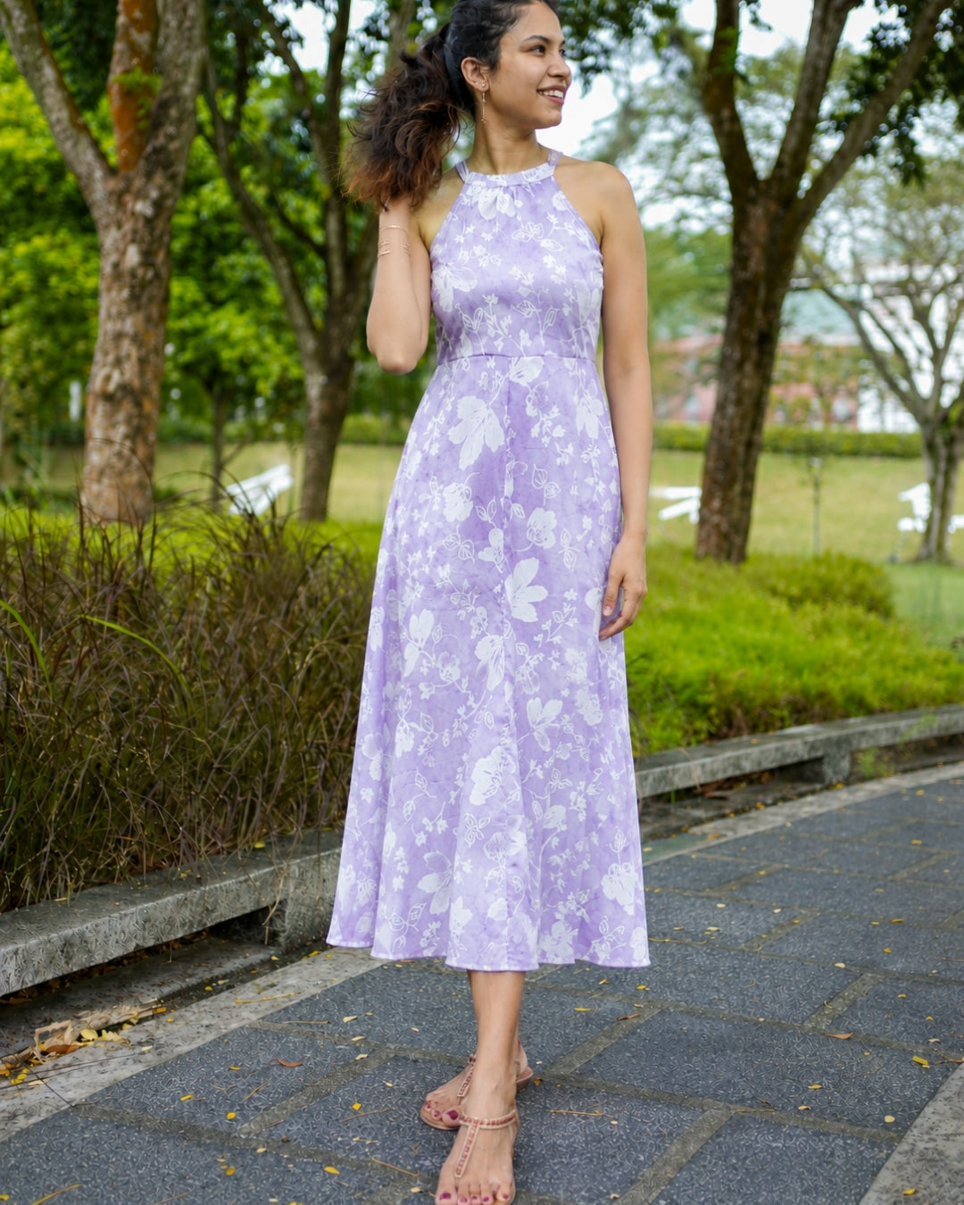 A woman wearing a sleeveless lavender halter dress with a white floral pattern, standing on a paved path in a park with trees in the background.
