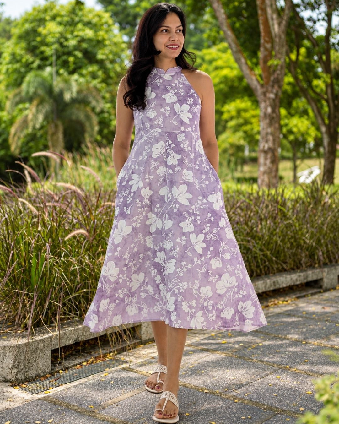 A woman wearing a sleeveless lavender halter dress with a white floral pattern, standing on a paved path in a park with trees in the background.