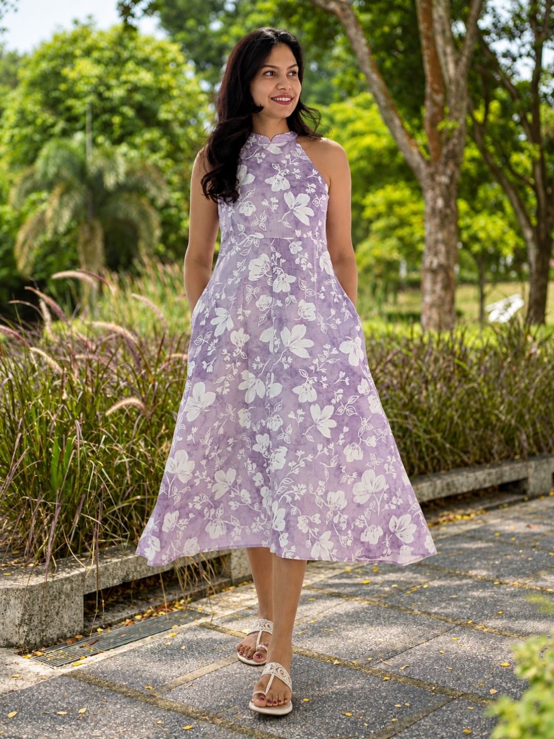 A woman wearing a sleeveless lavender halter dress with a white floral pattern, standing on a paved path in a park with trees in the background.