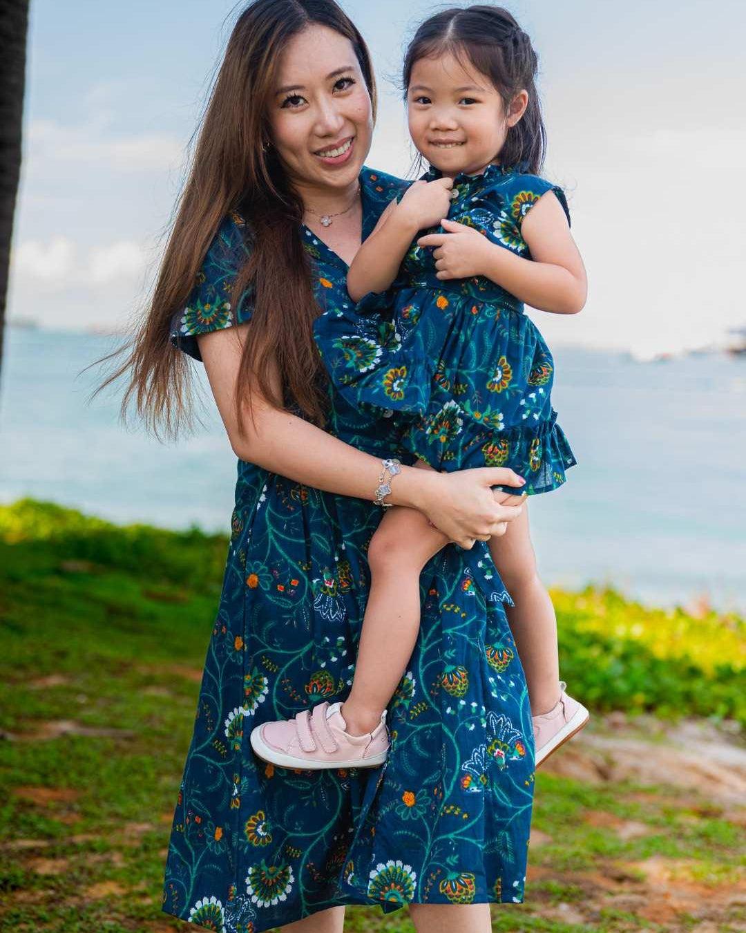 Woman holding a child both wearing matching blue floral dresses by the ocean.