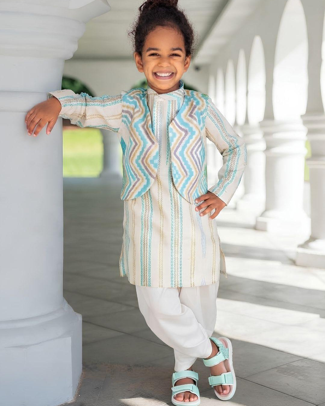 A young child wearing a traditional outfit with a striped kurta and a patterned jacket, standing in a sunny outdoor corridor with white pillars.