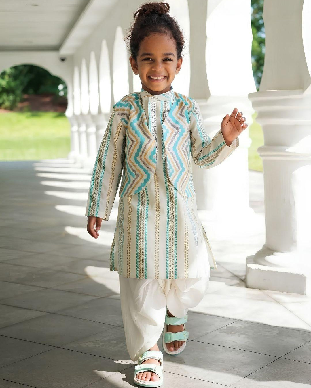 A young child walking on a covered walkway wearing a light beige traditional outfit with turquoise and white zigzag and vertical stripe patterns, paired with white pants and sandals.