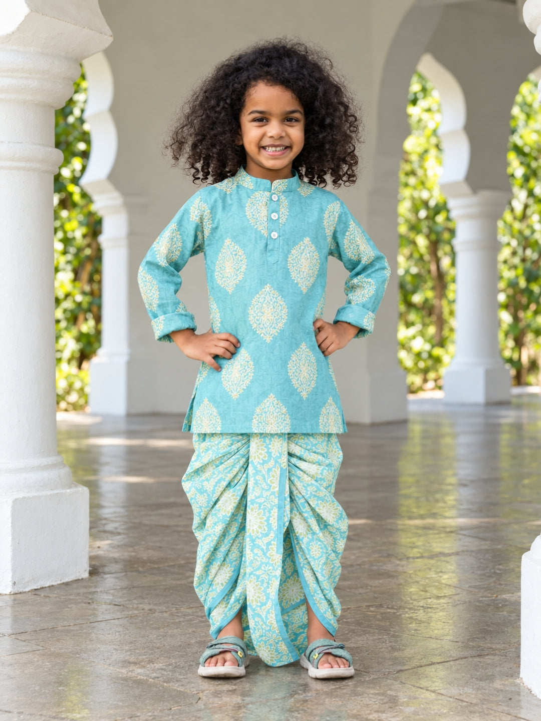 A young child with curly dark hair wearing a light turquoise blue and white printed traditional outfit, standing in front of white architectural columns.