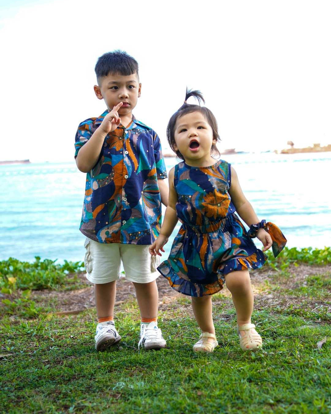 Two children in matching outfits standing on grass with a body of water in the background