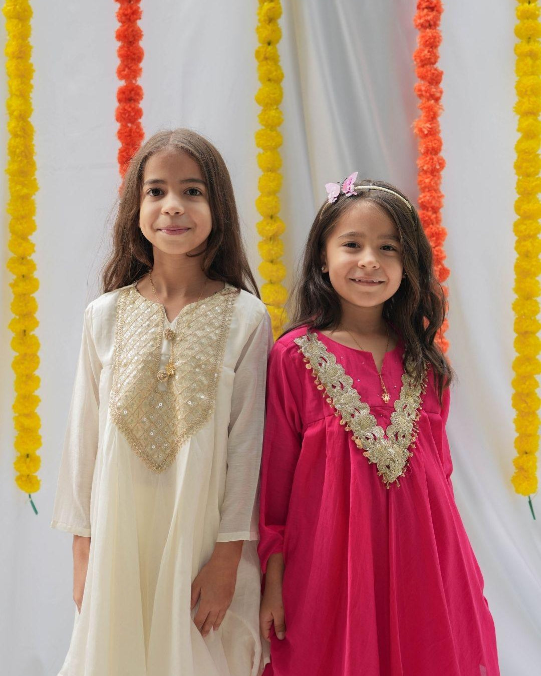 Two young girls standing side by side in traditional South Asian attire. The girl on the left wears an off-white kurta with a gold embroidered yoke, and the girl on the right wears a vibrant magenta-pink kurta with a matching gold embroidered yoke.