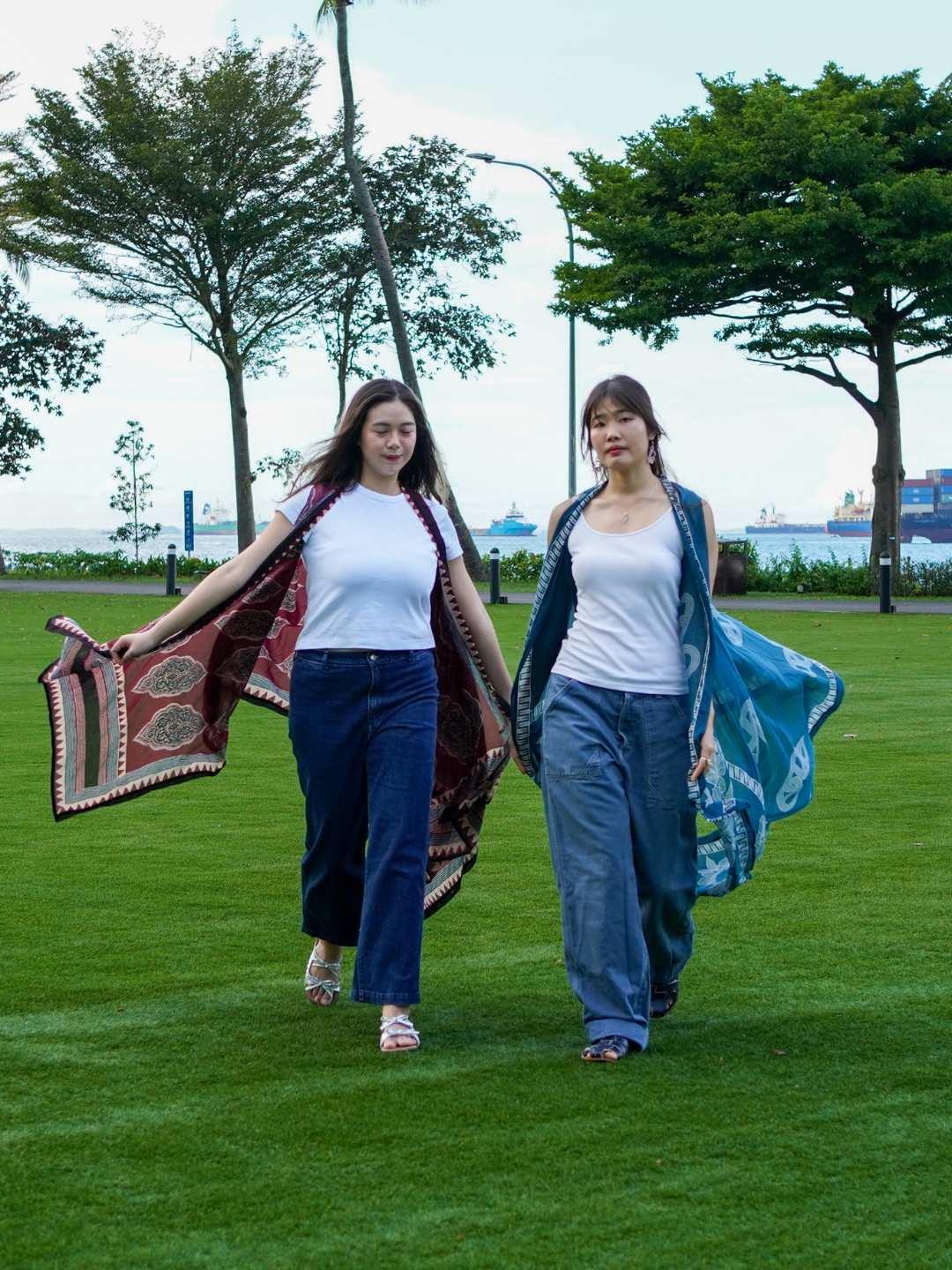 Two women walking on grass holding patterned cloths, with trees and a building in the background.