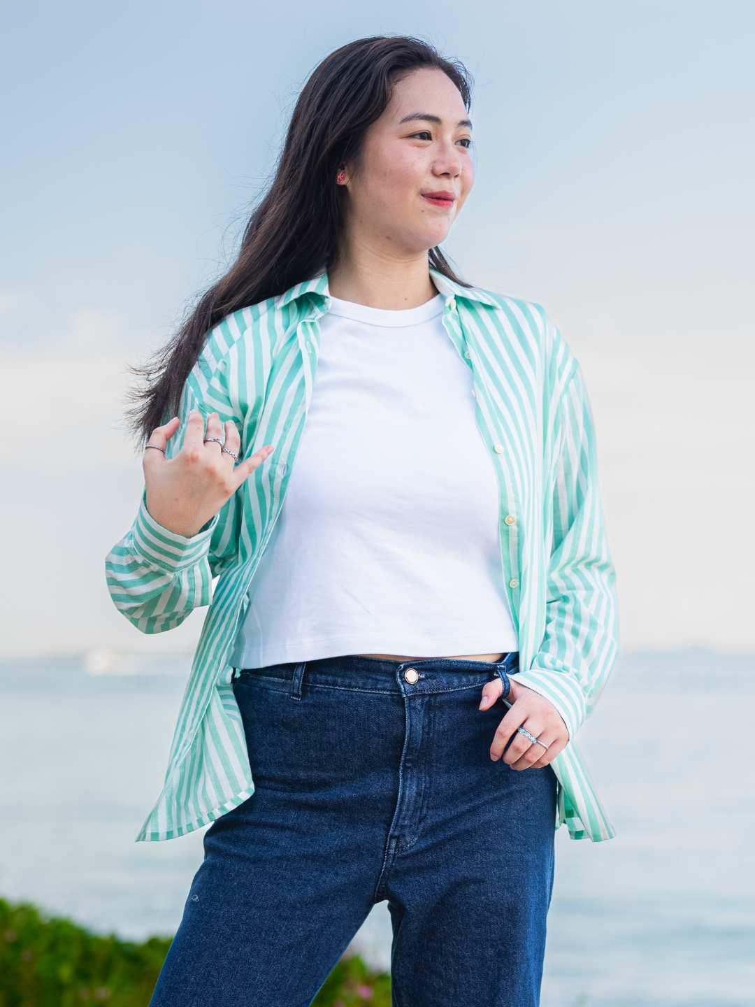 Woman wearing a green and white striped shirt over a white top with blue jeans, standing outdoors.