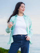 Woman wearing a green and white striped shirt over a white top with blue jeans, standing outdoors.
