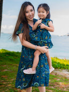 Woman holding a child both wearing matching blue floral dresses by the ocean.