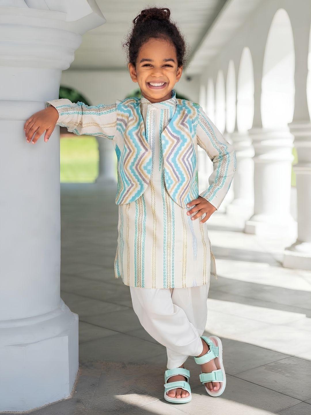 A young child wearing a traditional outfit with a striped kurta and a patterned jacket, standing in a sunny outdoor corridor with white pillars.