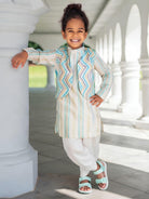 A young child wearing a traditional outfit with a striped kurta and a patterned jacket, standing in a sunny outdoor corridor with white pillars.