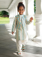A young child walking on a covered walkway wearing a light beige traditional outfit with turquoise and white zigzag and vertical stripe patterns, paired with white pants and sandals.