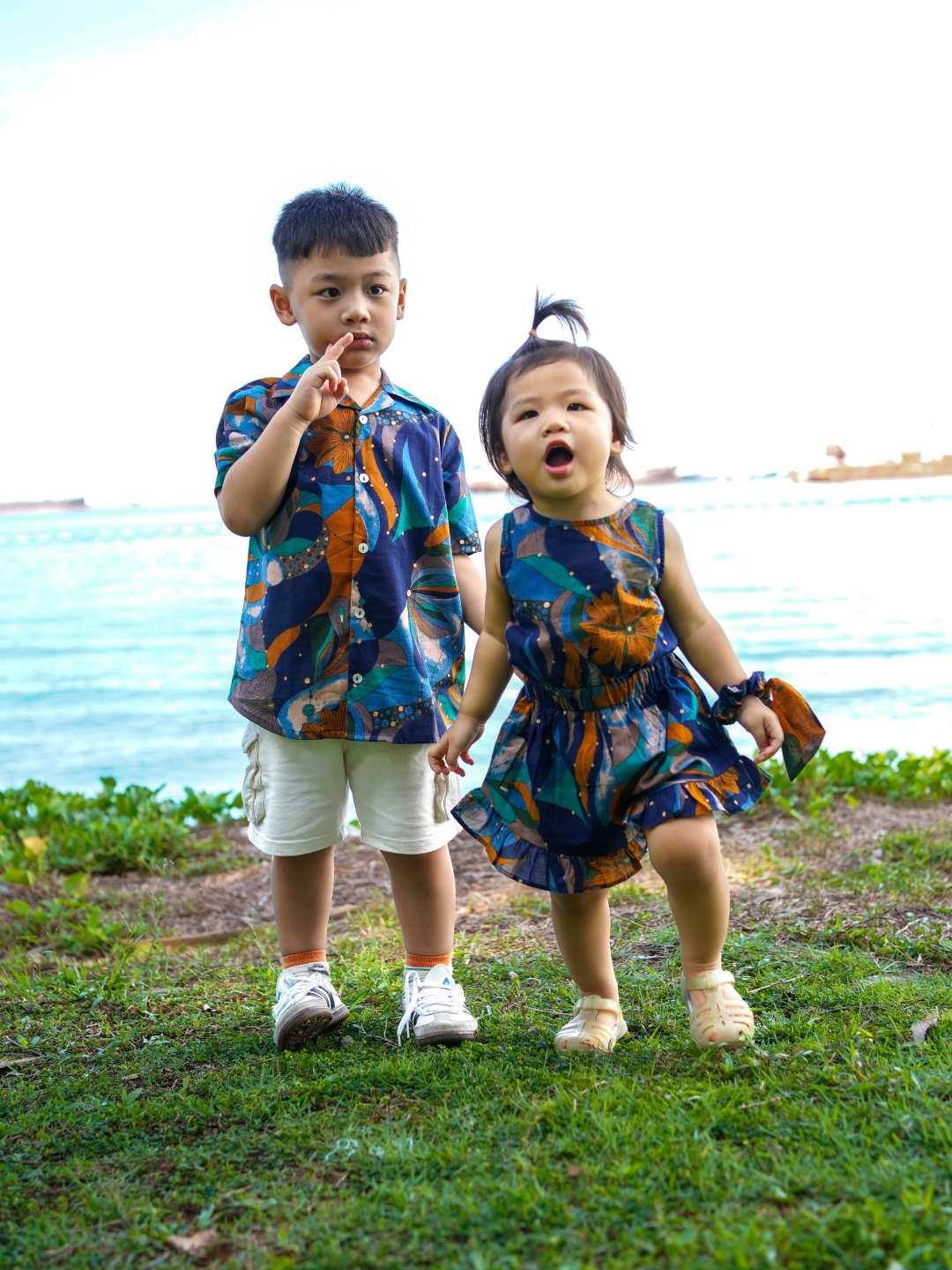 Two children in matching outfits standing on grass with a body of water in the background