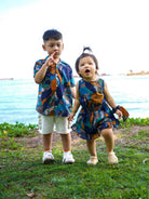 Two children in matching outfits standing on grass with a body of water in the background