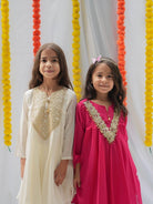 Two young girls standing side by side in traditional South Asian attire. The girl on the left wears an off-white kurta with a gold embroidered yoke, and the girl on the right wears a vibrant magenta-pink kurta with a matching gold embroidered yoke.