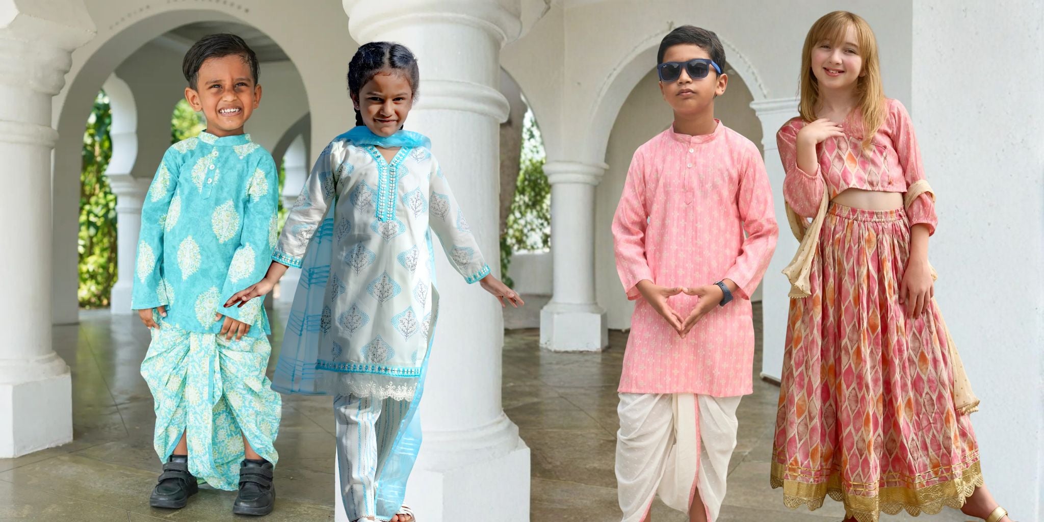 Four children modeling traditional Indian ethnic wear in a white arched corridor. The outfits include a light blue patterned kurta and dhoti, a white and light blue embroidered kurta and pants, a pink kurta with white dhoti, and a pink patterned kurta and matching skirt.