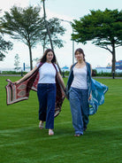 Two women walking on grass holding patterned cloths, with trees and a building in the background.