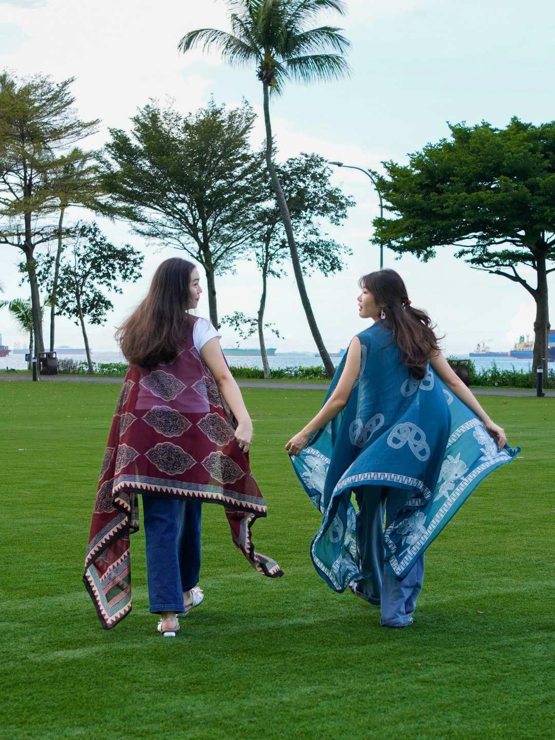 Two women walking on grass holding patterned cloths, with trees and water in the background.